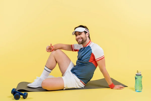 Joven deportista mostrando señal de paz mientras está sentado en la colchoneta de fitness cerca de pesas y botellas deportivas sobre fondo amarillo - foto de stock