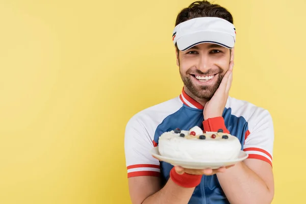 Позитивный спортсмен в козырьке держит размытый торт изолированный на желтый — стоковое фото