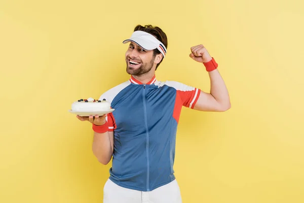 Young sportsman showing muscles and holding cake isolated on yellow — Stock Photo