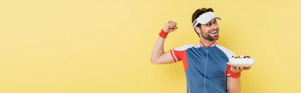 Cheerful sportsman holding cake and showing muscles isolated on yellow, banner — Stock Photo