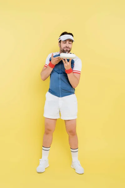 Full length of young sportsman looking at tasty cake on yellow background — Stock Photo