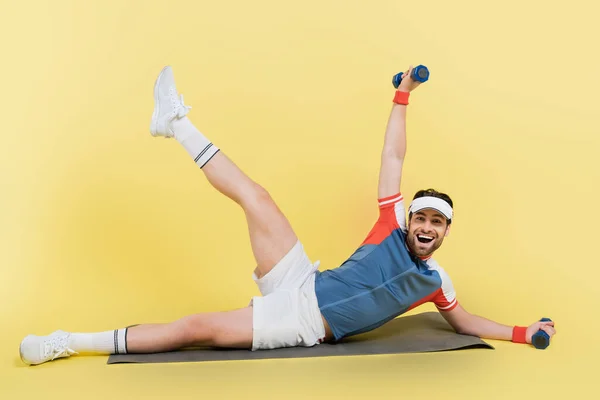 Positive sportsman holding dumbbells while training on fitness mat on yellow background — Stock Photo