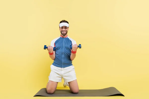 Esportista alegre segurando halteres e olhando para a câmera no tapete de fitness no fundo amarelo — Fotografia de Stock