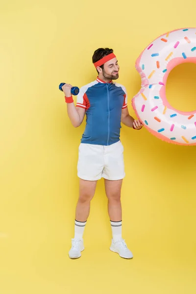 Pleine longueur de sportif souriant tenant anneau de natation et haltère sur fond jaune — Photo de stock