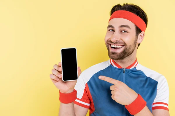Позитивный спортсмен указывает на смартфон с пустым экраном, изолированным на желтом — стоковое фото