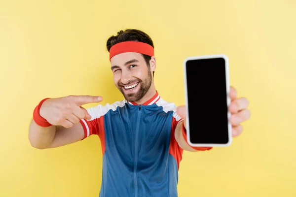 Усміхнений спортсмен підморгує і вказує на розмитий смартфон з порожнім екраном ізольовано на жовтому — стокове фото