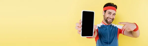 Deportista positivo guiñando un guiño y señalando el teléfono celular aislado en amarillo, banner - foto de stock