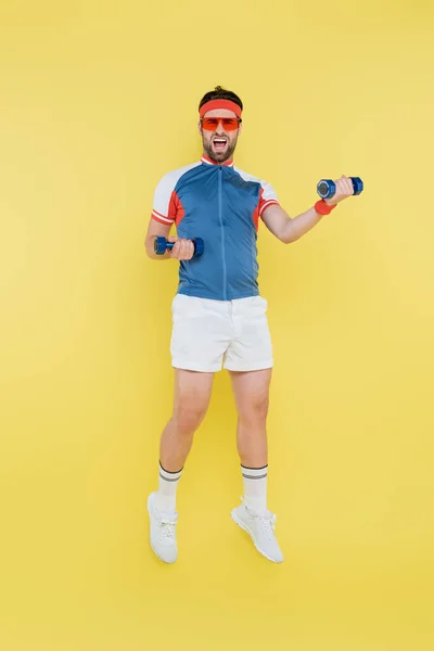 Sportsman in sunglasses jumping and holding dumbbells isolated on yellow — Stock Photo
