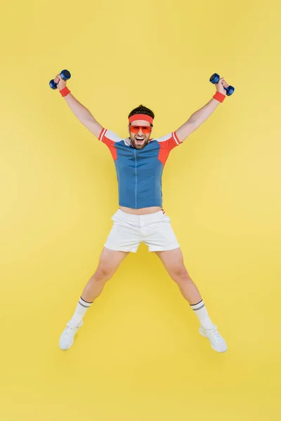Esportista positivo segurando halteres e pulando isolado no amarelo — Fotografia de Stock