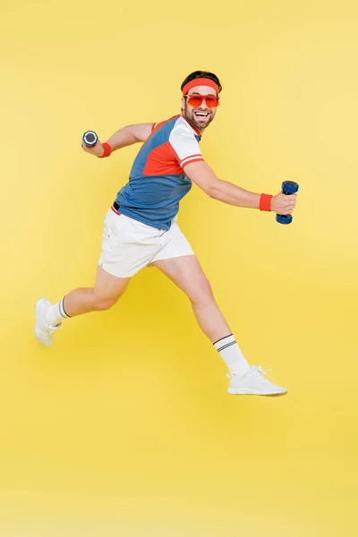 Deportista sonriente en gafas de sol saltando y sosteniendo pesas aisladas en amarillo - foto de stock