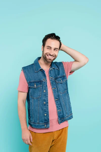 Smiling man in denim vest touching hair isolated on blue — Stock Photo