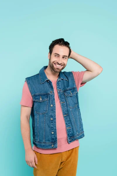 Uomo sorridente in gilet di denim toccare i capelli isolati su blu — Foto stock