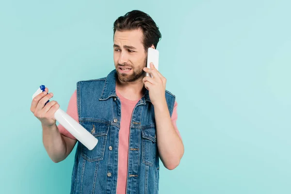 Confuso homem solteiro falando no smartphone e segurando detergente isolado no azul — Fotografia de Stock