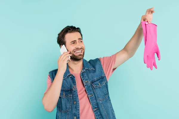 Homme souriant parlant sur téléphone portable et tenant des gants en caoutchouc isolés sur bleu — Photo de stock