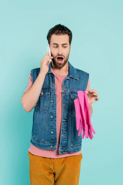 Shocked unmarried man looking at rubber gloves and talking on smartphone isolated on blue — Stock Photo