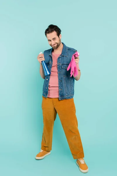 Pleine longueur d'homme souriant tenant des gants en caoutchouc et du détergent sur fond bleu — Photo de stock