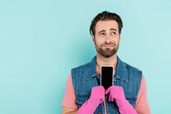 Hombre disgustado en guantes de goma que sostiene el teléfono celular aislado en azul - foto de stock