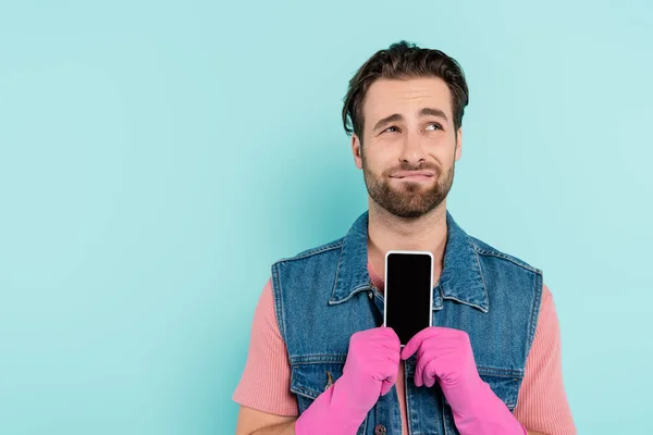 Displeased man in rubber gloves holding cellphone isolated on blue — Stock Photo