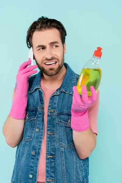 Escéptico hombre soltero en guantes de goma sosteniendo líquido lavavajillas y hablando en el teléfono inteligente aislado en azul - foto de stock
