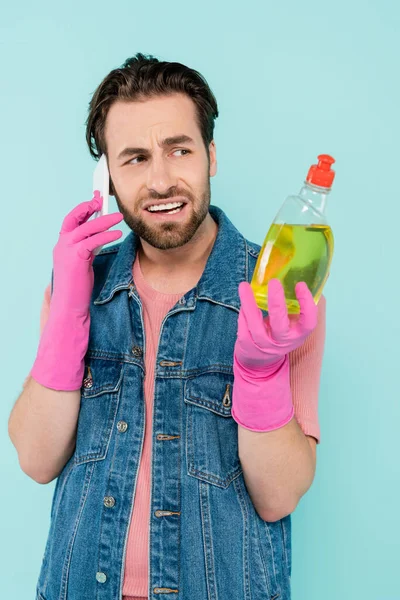 Skeptischer unverheirateter Mann in Gummihandschuhen hält Spülmittel in der Hand und spricht auf dem Smartphone isoliert auf blauem Grund — Stockfoto