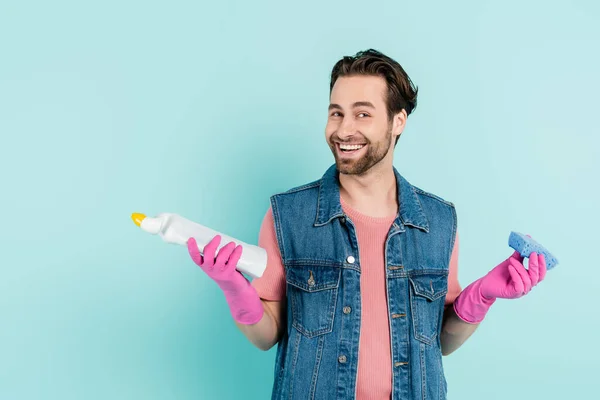 Positive young man in rubber gloves holding sponge and detergent isolated on blue — Stock Photo