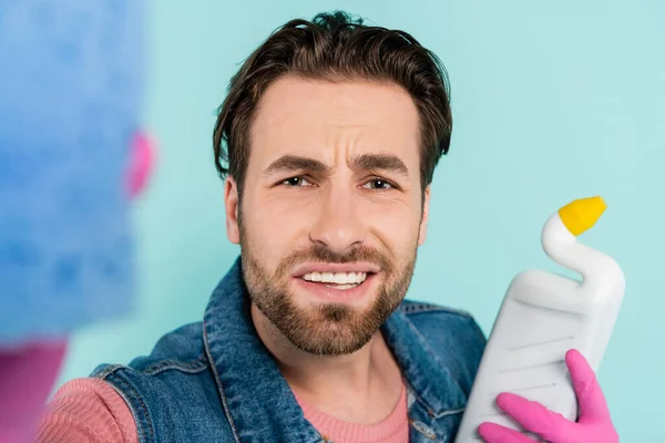 Joven con guantes de goma sujetando esponja y detergente aislado en azul - foto de stock