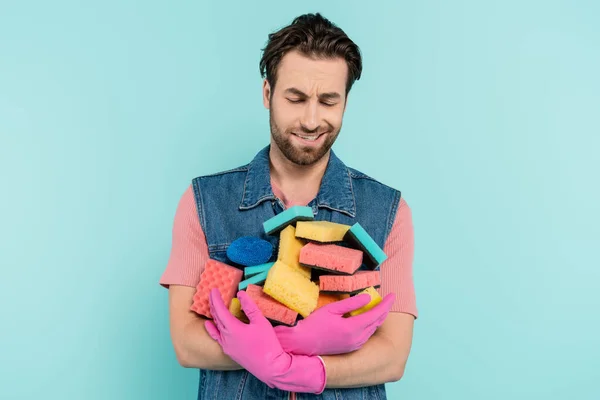 Hombre no casado disgustado con guantes de goma sosteniendo esponjas aisladas en azul - foto de stock