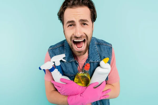 Joven hombre soltero con guantes de goma sosteniendo detergentes y gritando aislado en azul - foto de stock