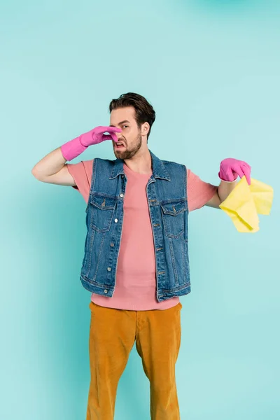 Young man in rubber gloves covering nose while holding rug isolated on blue — Stock Photo