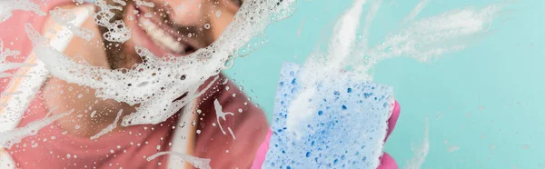 Vista ritagliata dell'uomo sorridente che pulisce vetro con detergente e spugna isolati su blu, banner — Foto stock