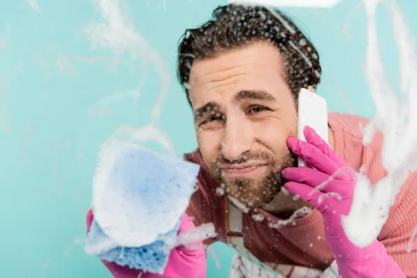 Focused unmarried man in rubber gloves talking on smartphone and cleaning glass isolated on blue — Stock Photo