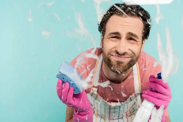 Hombre molesto en guantes de goma que sostienen detergente y esponja cerca de vidrio aislado en azul - foto de stock
