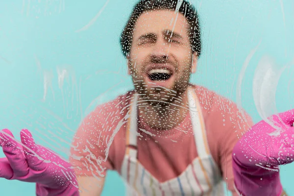 Hombre molesto borroso en guantes de goma de pie cerca de vidrio con detergente aislado en azul - foto de stock