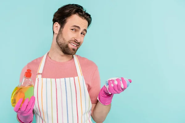 Sad man in apron and rubber gloves holding dishwashing liquid and sponge isolated on blue — Stock Photo