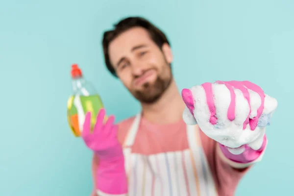 Homme flou en gants de caoutchouc tenant détergent et éponge avec mousse isolée sur bleu — Photo de stock