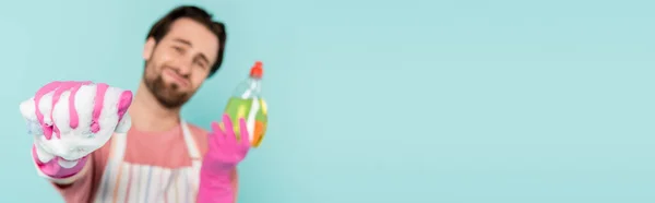 Blurred man in apron and rubber gloves holding detergent and sponge isolated on blue, banner — Stock Photo