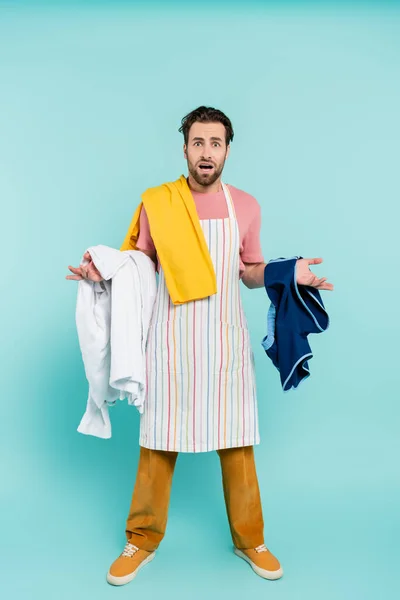 Confundido hombre soltero en delantal sosteniendo la ropa sobre fondo azul - foto de stock