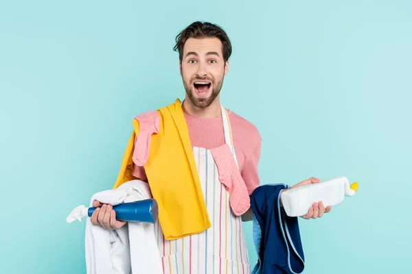 Aufgeregter Mann in Schürze hält Waschmittel und Kleidung isoliert auf blauem Grund — Stockfoto