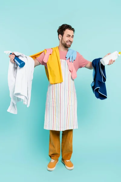 Comprimento total do homem sorridente em avental segurando detergentes e roupas em fundo azul — Fotografia de Stock