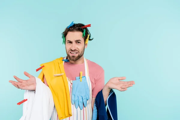 Displeased unmarried man in clothespins holding clothes isolated on blue — Stock Photo