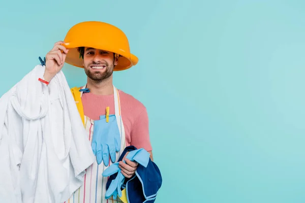 Cheerful man in clothespins holding wash bowl on head and clothes isolated on blue — Stock Photo