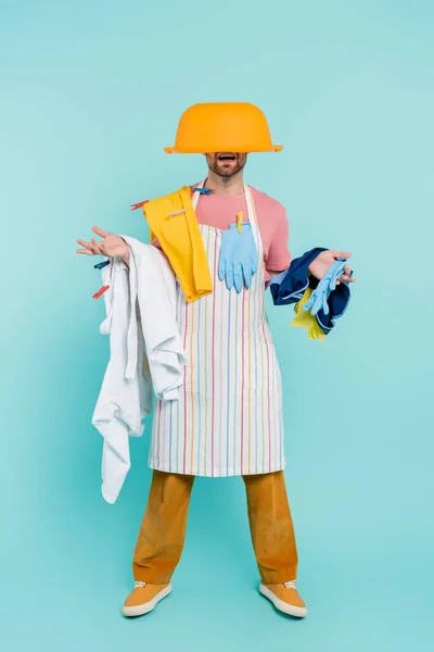 Shocked unmarried man in clothespins, holding clothes and wash bowl on head on blue background — Stock Photo