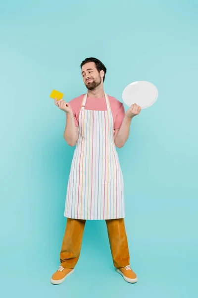 Full length of tired unmarried man in apron holding sponge and plate on blue background — Stock Photo