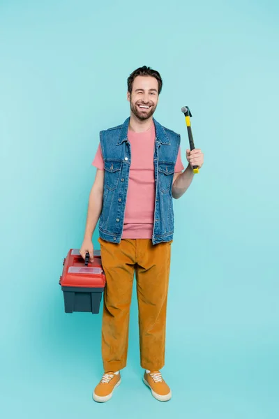 Full length of positive man holding hammer and toolbox on blue background — Stock Photo