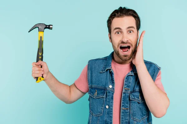 Shocked man in denim vest holding hammer isolated on blue — Stock Photo