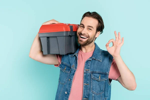 Fröhlicher Mann in Jeansweste hält Werkzeugkiste in der Hand und zeigt OK-Geste isoliert auf blau — Stockfoto