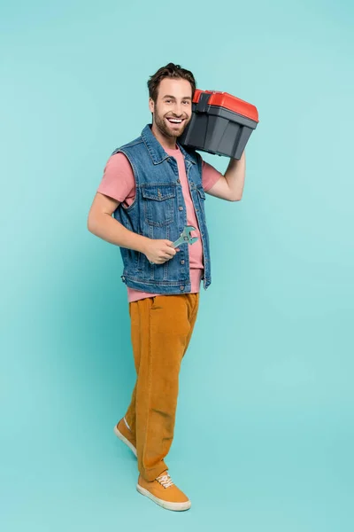 Longitud completa del hombre positivo sosteniendo la llave y la caja de herramientas sobre fondo azul - foto de stock