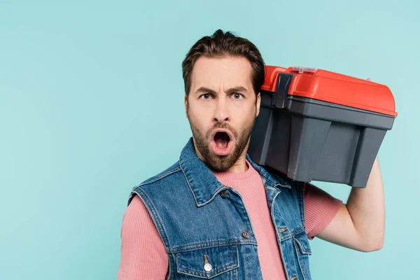Young shocked man looking at camera and holding toolbox isolated on blue — Stock Photo