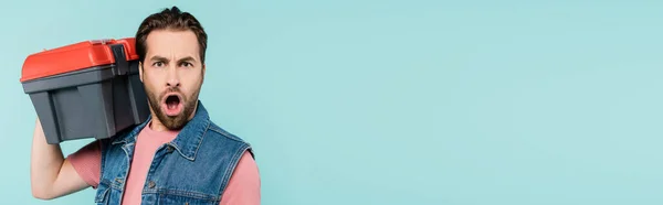 Shocked man looking at camera while holding toolbox isolated on blue, banner — Stock Photo