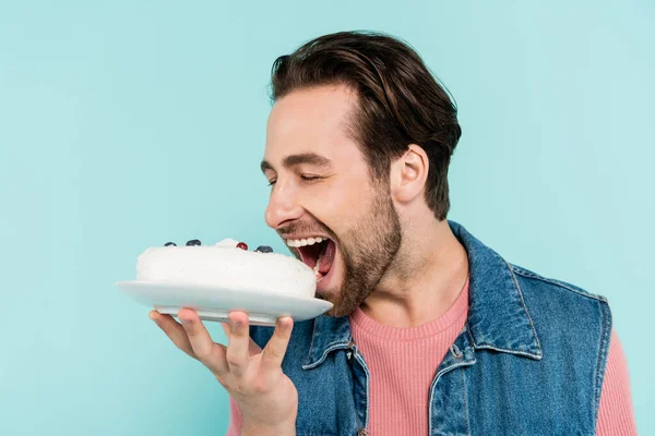 Joven con chaleco vaquero abriendo la boca cerca de la torta aislado en azul - foto de stock